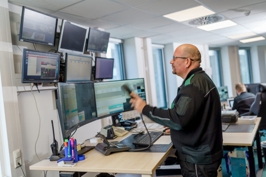 Ein Mann in dunkler Arbeitskleidung steht in einer Leitstelle vor mehreren Monitoren und hält einen Telefonhörer in der Hand.