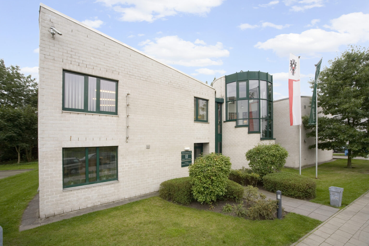 Modernes Bürogebäude aus hellen Ziegeln mit grünen Fensterrahmen, einem gläsernen Treppenhaus und Flaggen vor dem Eingangsbereich.