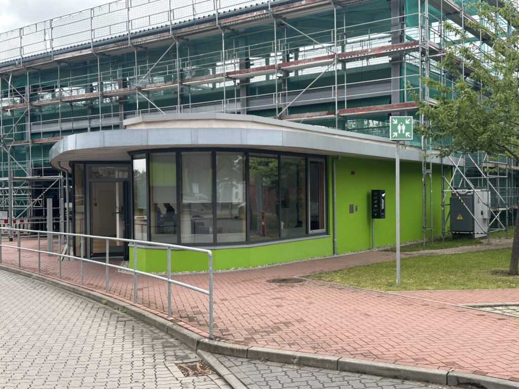 Ein flaches, hellgrünes Gebäude mit großen Glasfronten vor einem mit Gerüst und grünen Netzen verkleideten Rohbau.