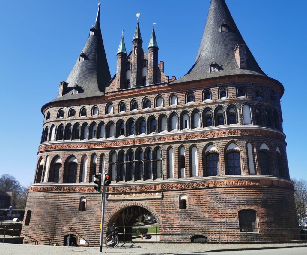 Historisches Stadttor aus Backstein mit zwei großen Rundtürmen, zahlreichen Fensterbögen und einer zentralen Durchfahrt vor blauem Himmel.