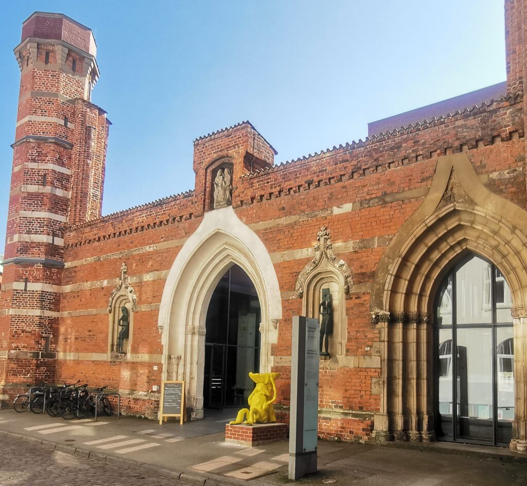 Historisches Backsteingebäude mit spitzbogigen Eingängen, einem Turm, Statuen in Nischen und einer gelben Skulptur im Vordergrund.
