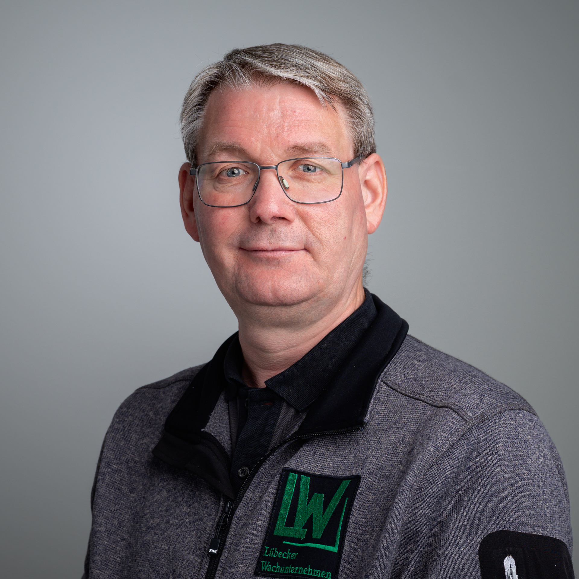 Porträt von Daniel Riemann, einem Mann mit Brille und grauem Haar, der eine graue Jacke mit dem grünen Logo des Lübecker Wachunternehmens trägt.