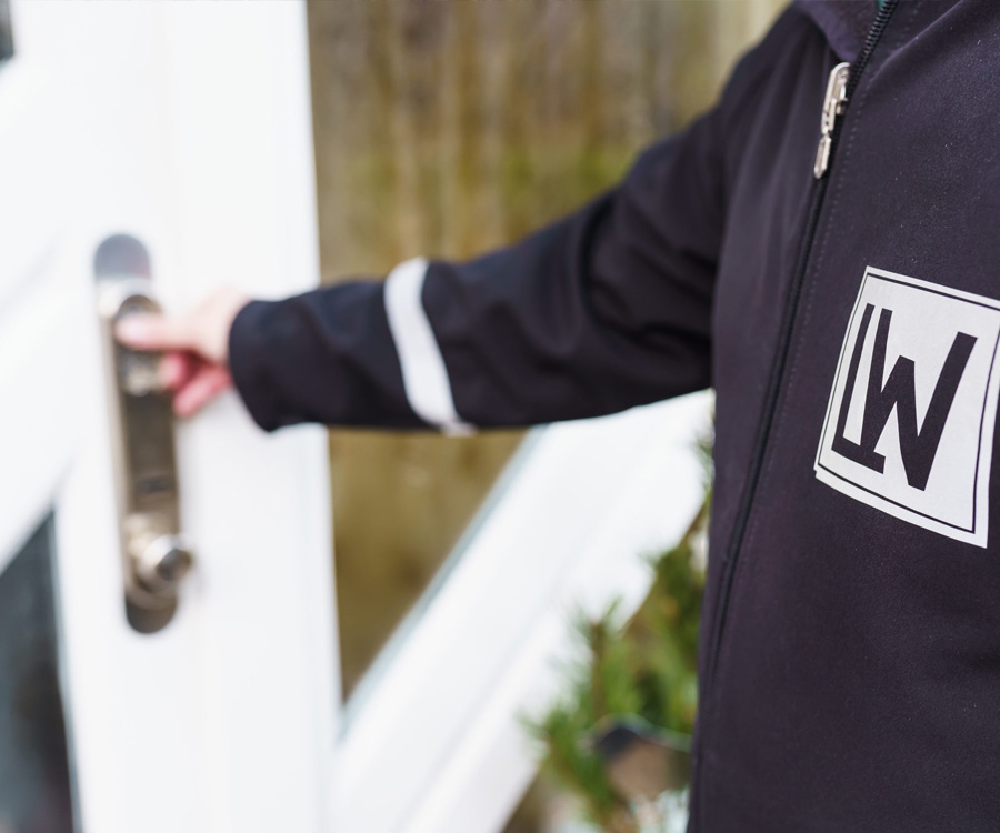Eine Person in einer dunklen Jacke mit Firmenlogo öffnet eine weiße Tür, eventuell im Rahmen einer Alarmaufschaltung.