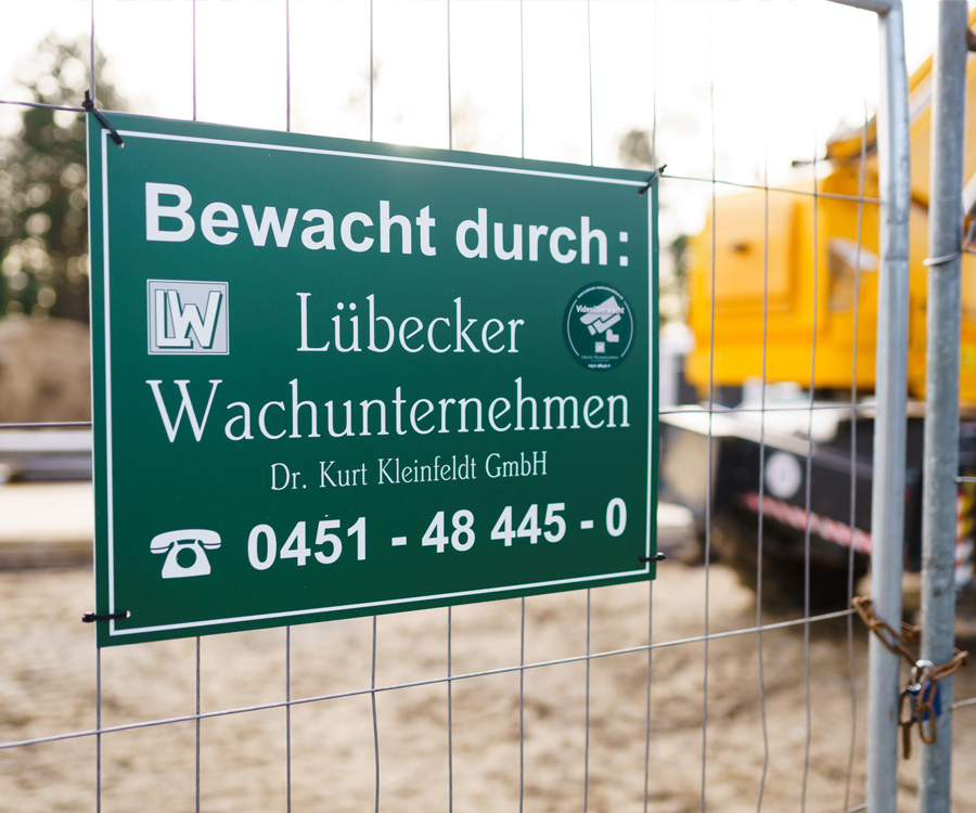 Ein grünes Schild an einem Bauzaun weist auf die Bewachung durch das Lübecker Wachunternehmen Dr. Kurt Kleinfeldt GmbH hin, inklusive Telefonnummer und Videoübe