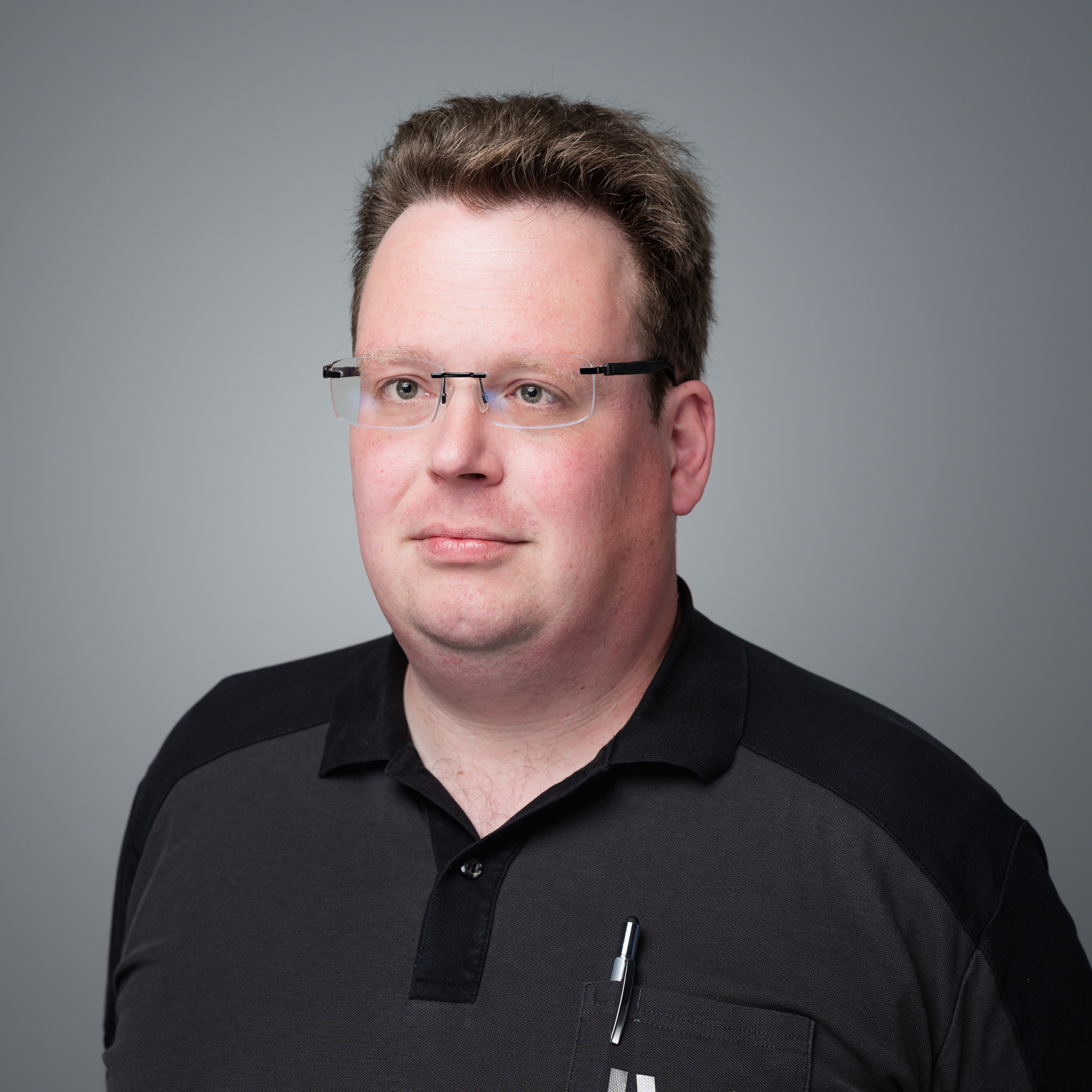 Porträtfoto von Dennis Saarloos, einem Mann mit Brille und dunklem Polo-Shirt, der neutral vor grauem Hintergrund leicht zur Seite blickt.