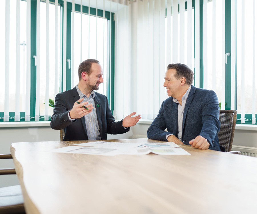 Zwei Männer besprechen lächelnd eine Gefahrenanalyse an einem Holztisch im Büro, wobei der eine gestikuliert und der andere aufmerksam zuhört.