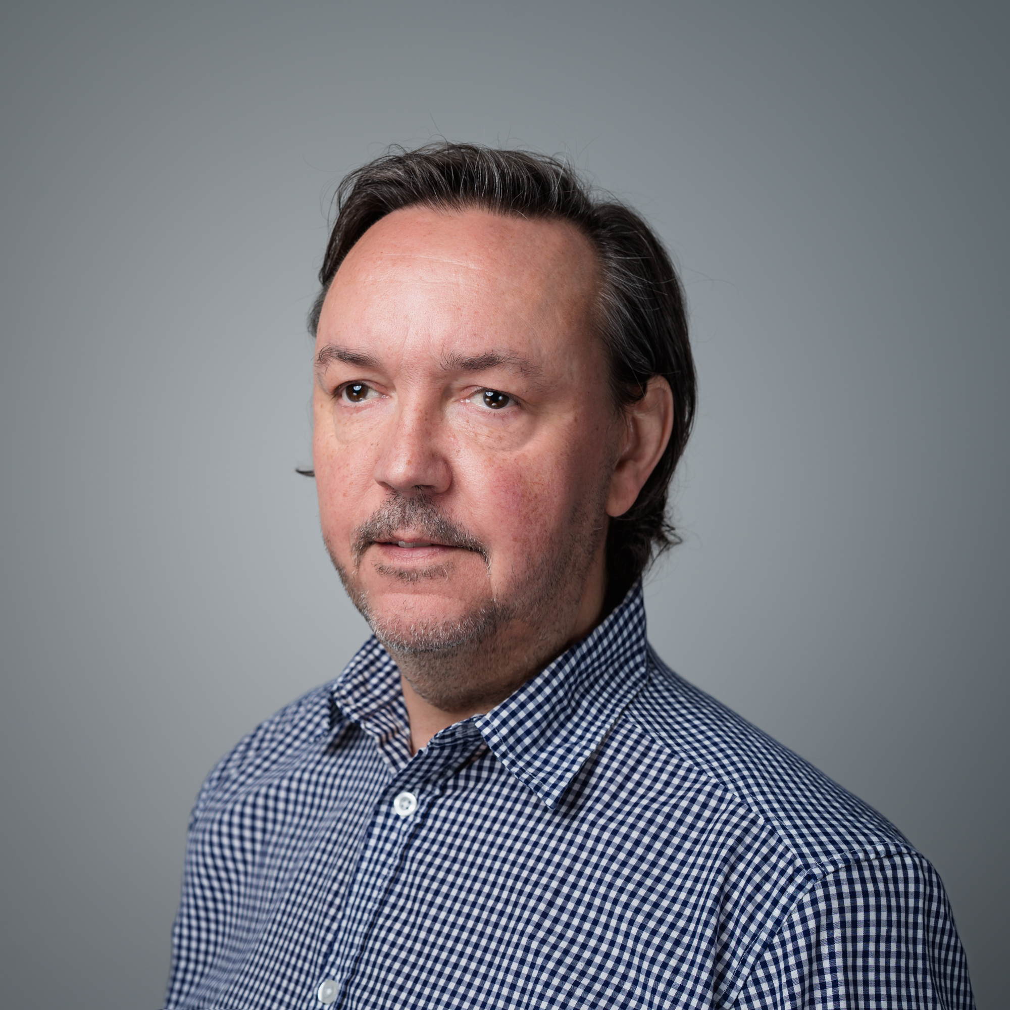 Porträt von Gerd Grzywatz, einem Mann mittleren Alters mit dunklem Haar und Bart, der ein blau-weiß kariertes Hemd trägt und zur Seite blickt.
