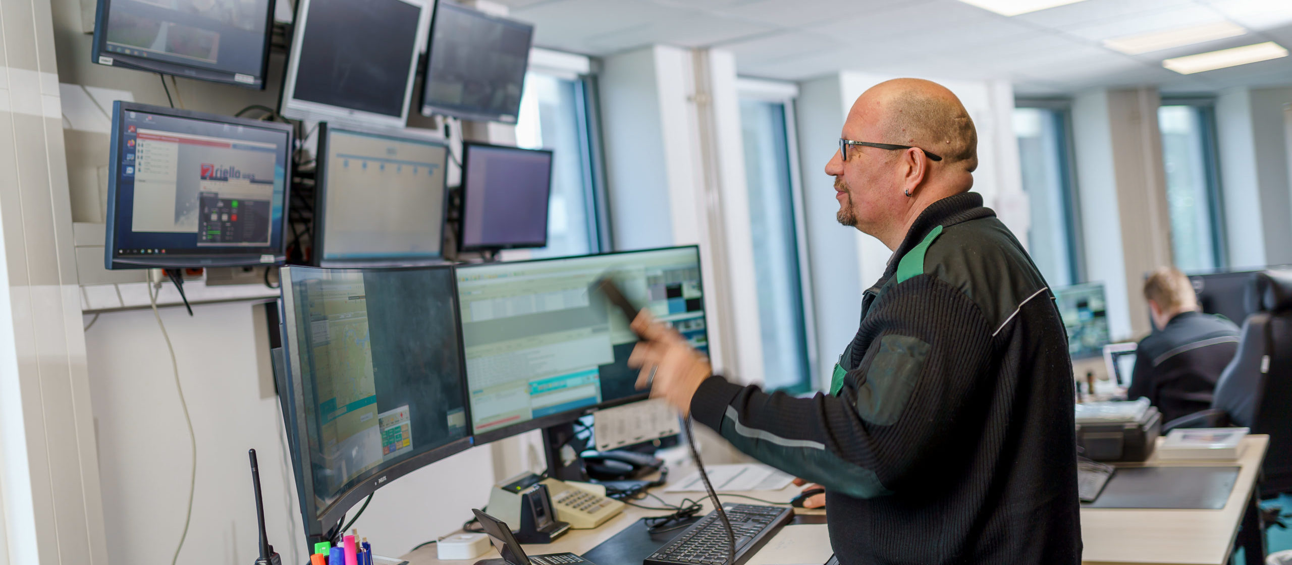 Ein Mann mit Brille und Bart steht vor mehreren Computermonitoren in einem Kontrollraum und bedient ein Gerät.