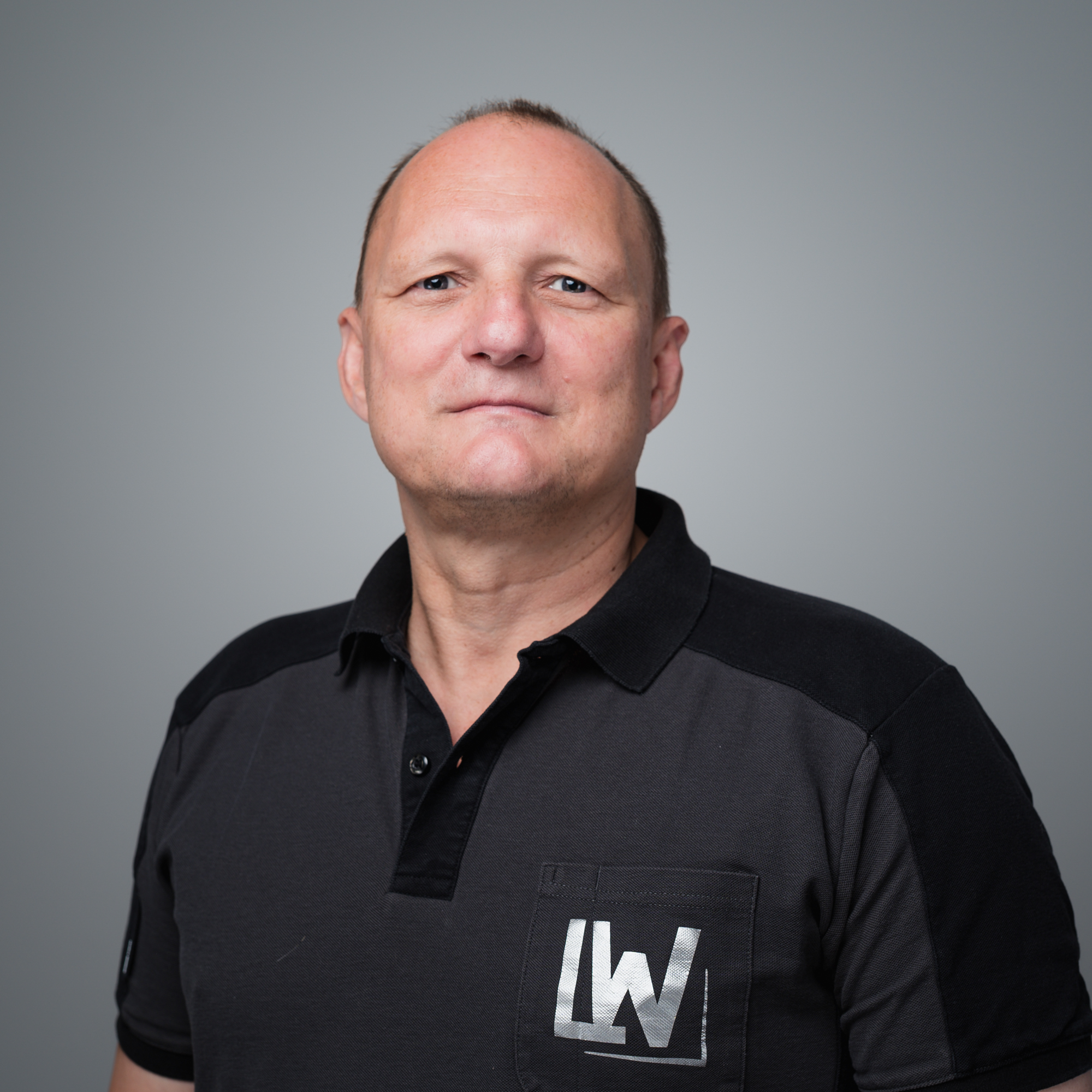 Porträt von Stefan Franck, einem Mann mittleren Alters mit dunklem Polo-Shirt und einem IW-Logo auf der Brusttasche, vor grauem Hintergrund.