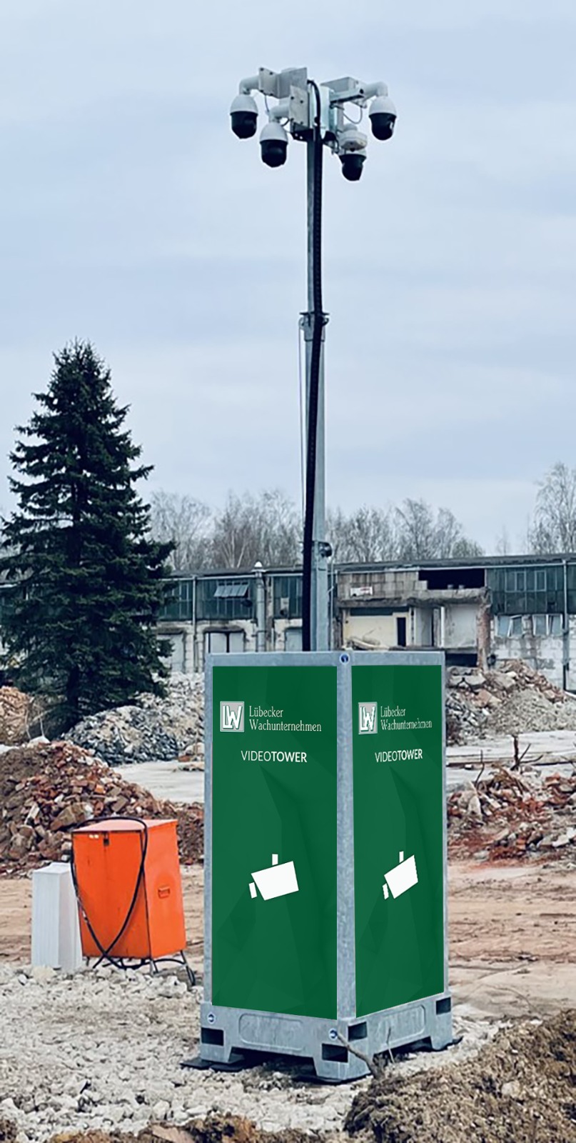 Ein grüner mobiler Überwachungsturm mit mehreren Kameras steht auf einem Schotterfeld vor einem teilweise abgerissenen Gebäude und einem großen Nadelbaum.