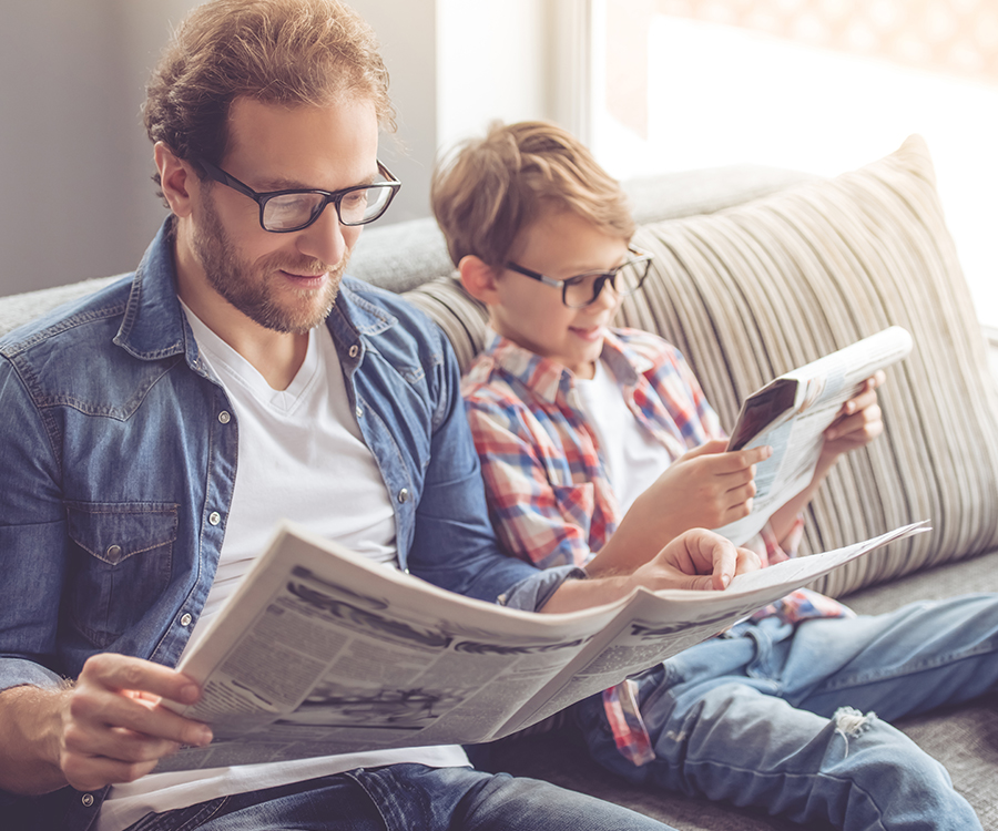 Ein Vater und sein Sohn sitzen auf einem Sofa und lesen gemeinsam aktuelle Nachrichten in Zeitungen, beide tragen Brillen.