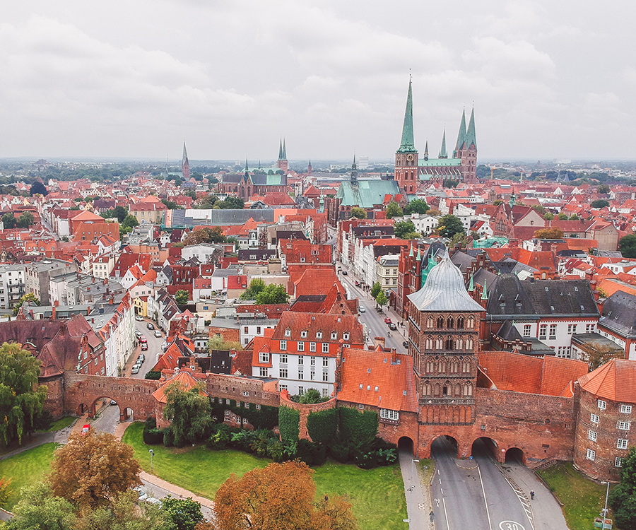 Luftaufnahme der historischen Altstadt von Lübeck mit dem Holstentor im Vordergrund und den markanten Kirchtürmen im Hintergrund unter bewölktem Himmel.
