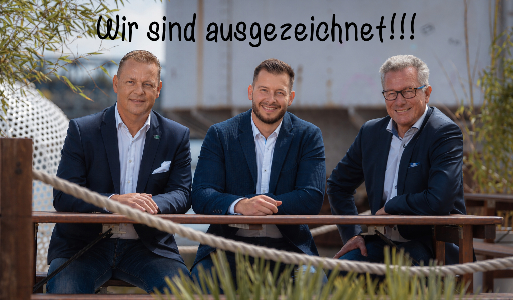 Drei lächelnde Männer in Anzügen sitzen an einem Holztisch im Freien, mit dem Text "Wir sind ausgezeichnet!!!" über dem Bild.