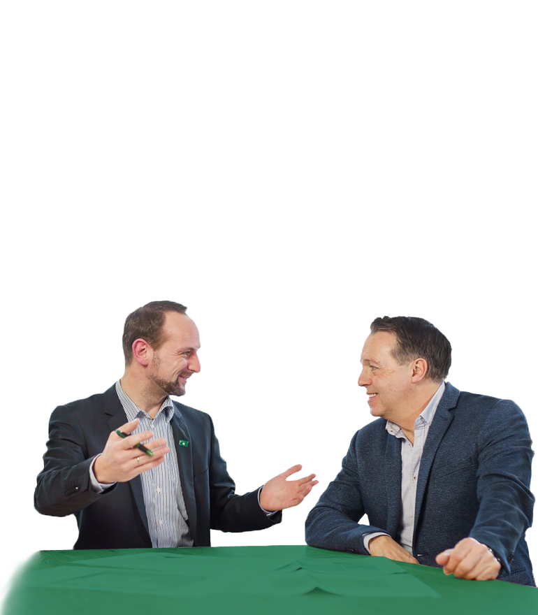 Zwei Männer in Business-Casual-Kleidung sitzen an einem grünen Tisch, lächeln und führen ein lebhaftes Gespräch.