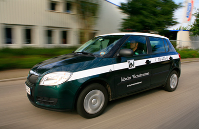 Ein dunkelgrünes Auto des Lübecker Wachunternehmens mit weißem Streifen fährt auf einer Straße, der Hintergrund ist durch die Bewegung verschwommen.
