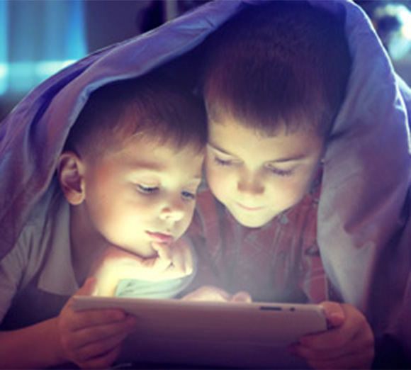 Zwei Jungen liegen unter einer Decke und schauen auf ein leuchtendes Tablet, dessen Licht ihre Gesichter erhellt.
